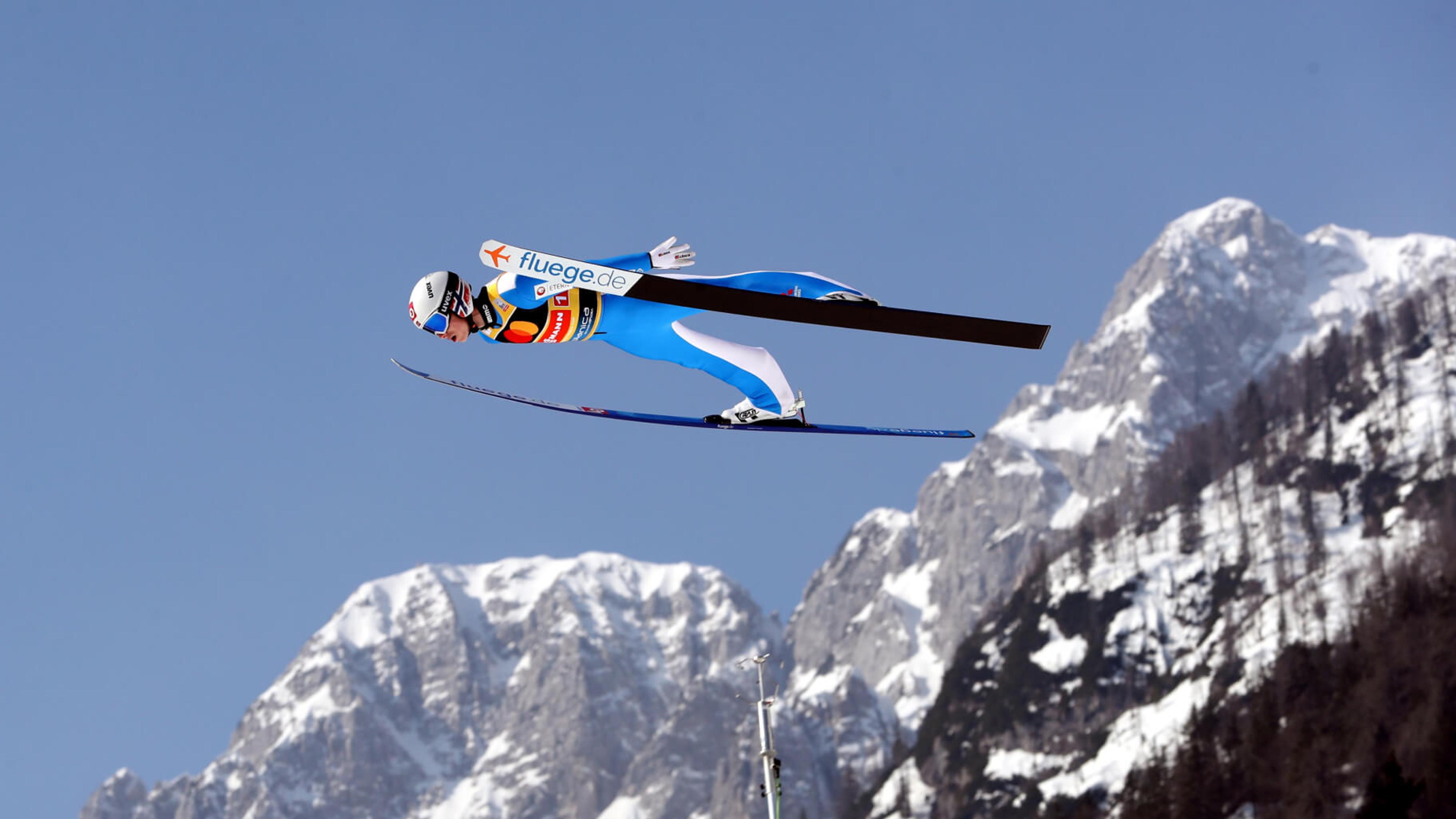 Skihopper svever høyt med blå himmel og snøkledde fjelltopper i bakgrunnen.