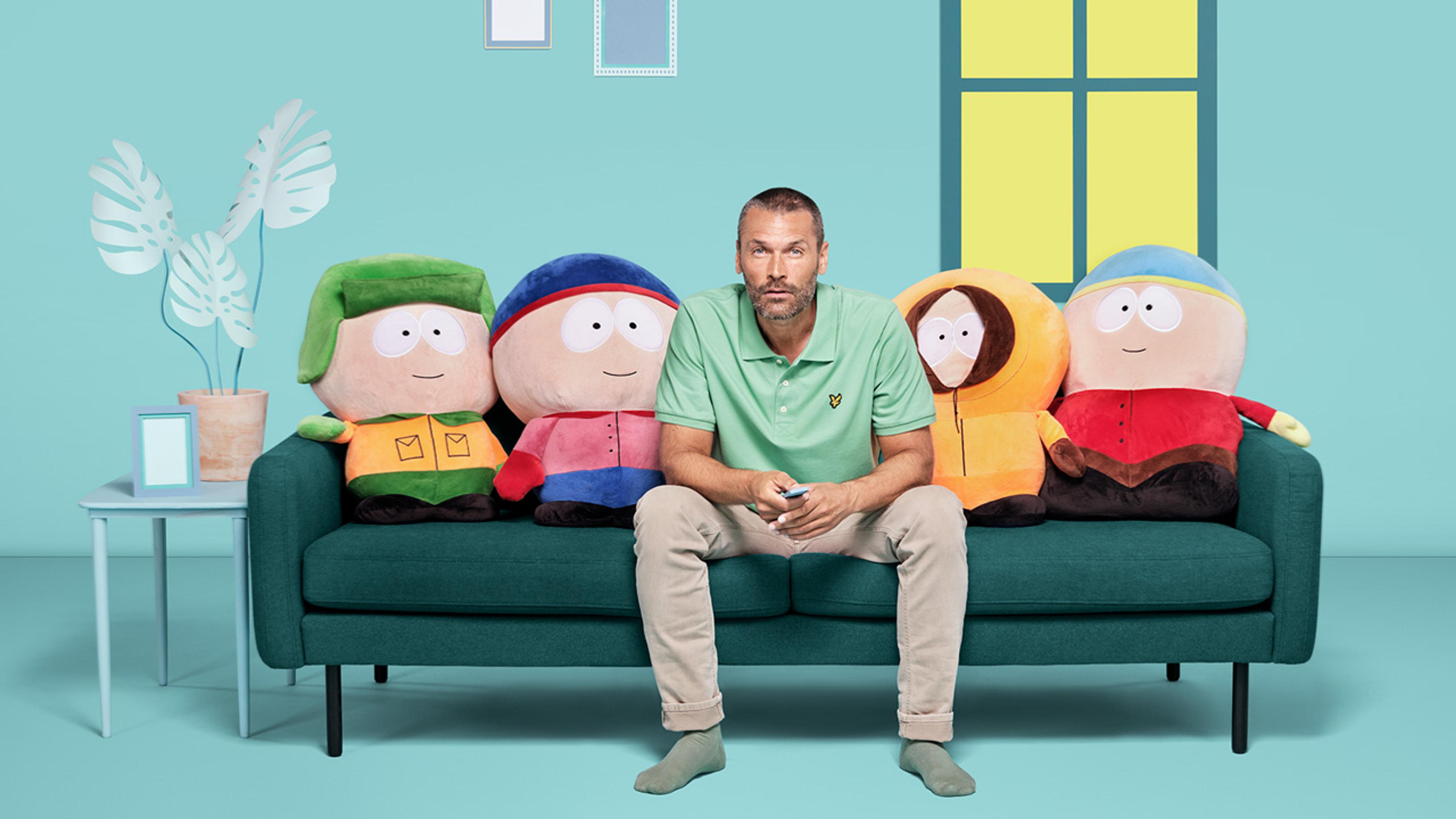Mann ser TV med Strim på en sofa full av South Park kosedyr.