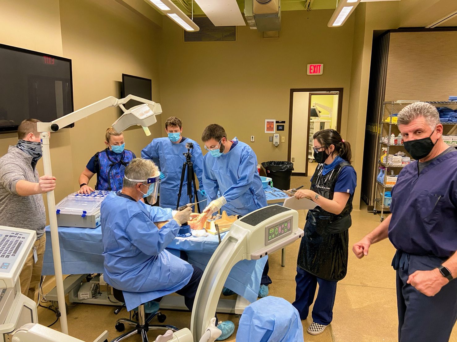 Medical team in surgical scrubs and masks performing a procedure in a clinical training room, gathered around a patient on an operating table with monitoring equipment and overhead lights, while another staff member stands nearby observing.