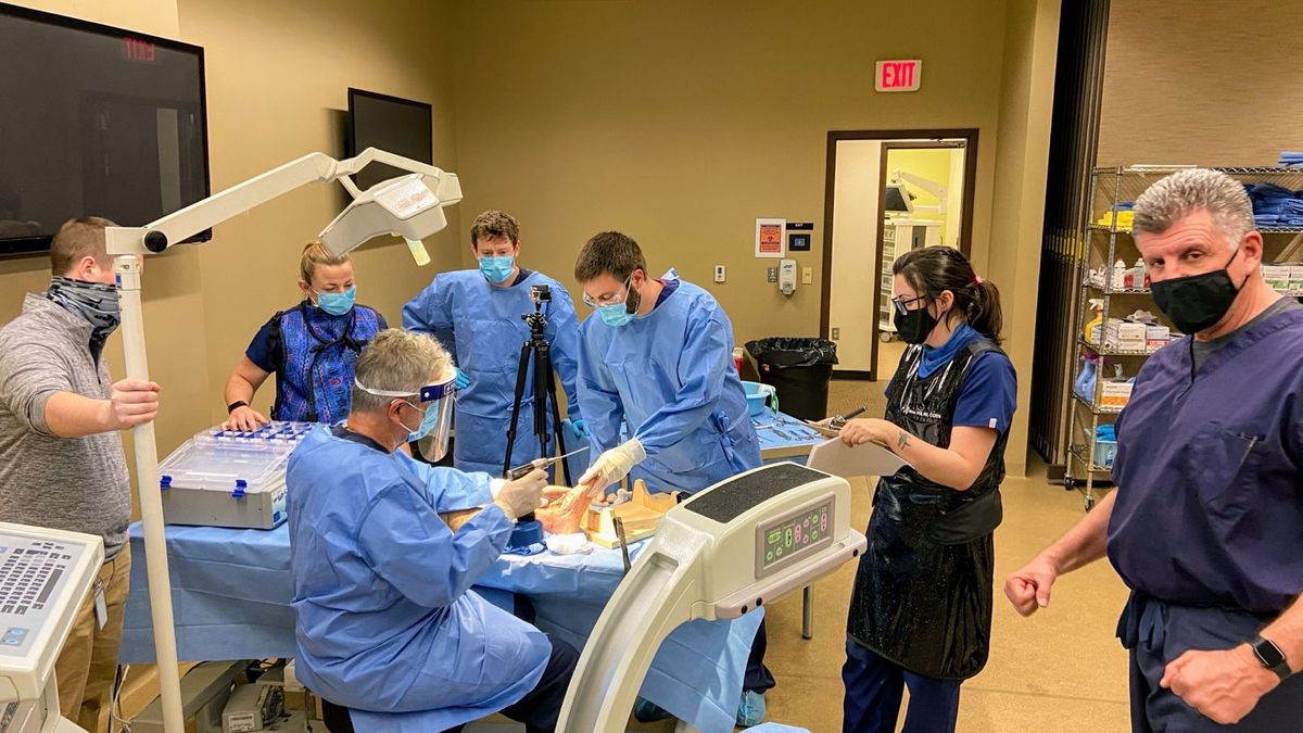 Medical team in surgical scrubs and masks performing a procedure in a clinical training room, gathered around a patient on an operating table with monitoring equipment and overhead lights, while another staff member stands nearby observing.
