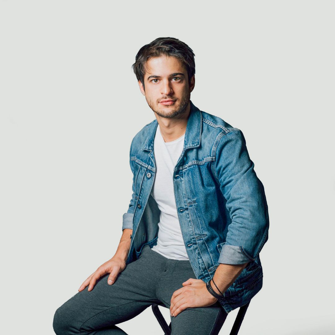 A studio portrait of a person with short brown hair sitting on a chair against a light grey background. The person wears a blue denim jacket over a white crew-neck t-shirt, dark charcoal trousers, and black bracelets on one wrist, looking straight towards the camera with hands resting on thighs.