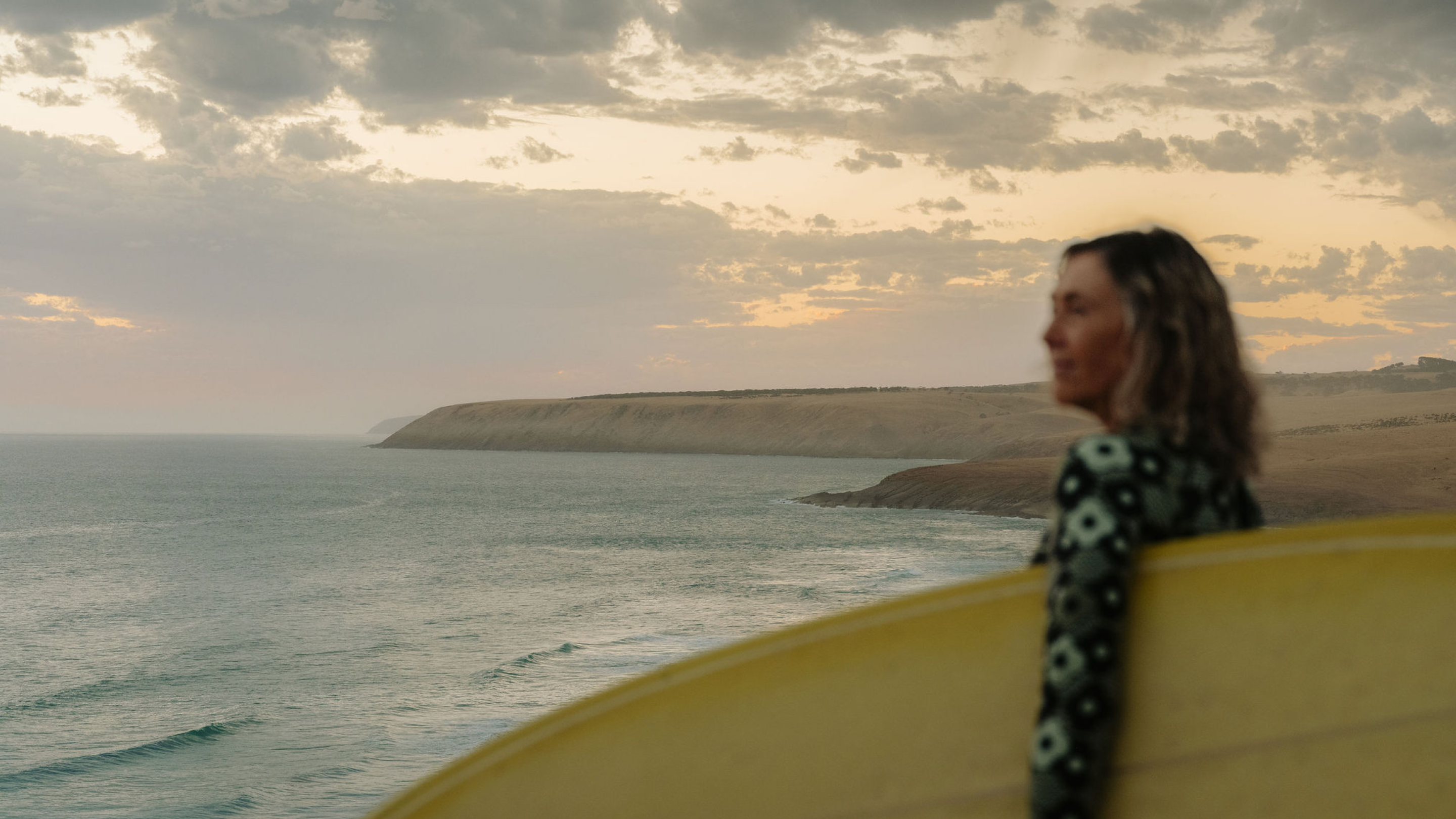 Surfer carrying a yellow surfboard beside the South Australian coast at sunset.