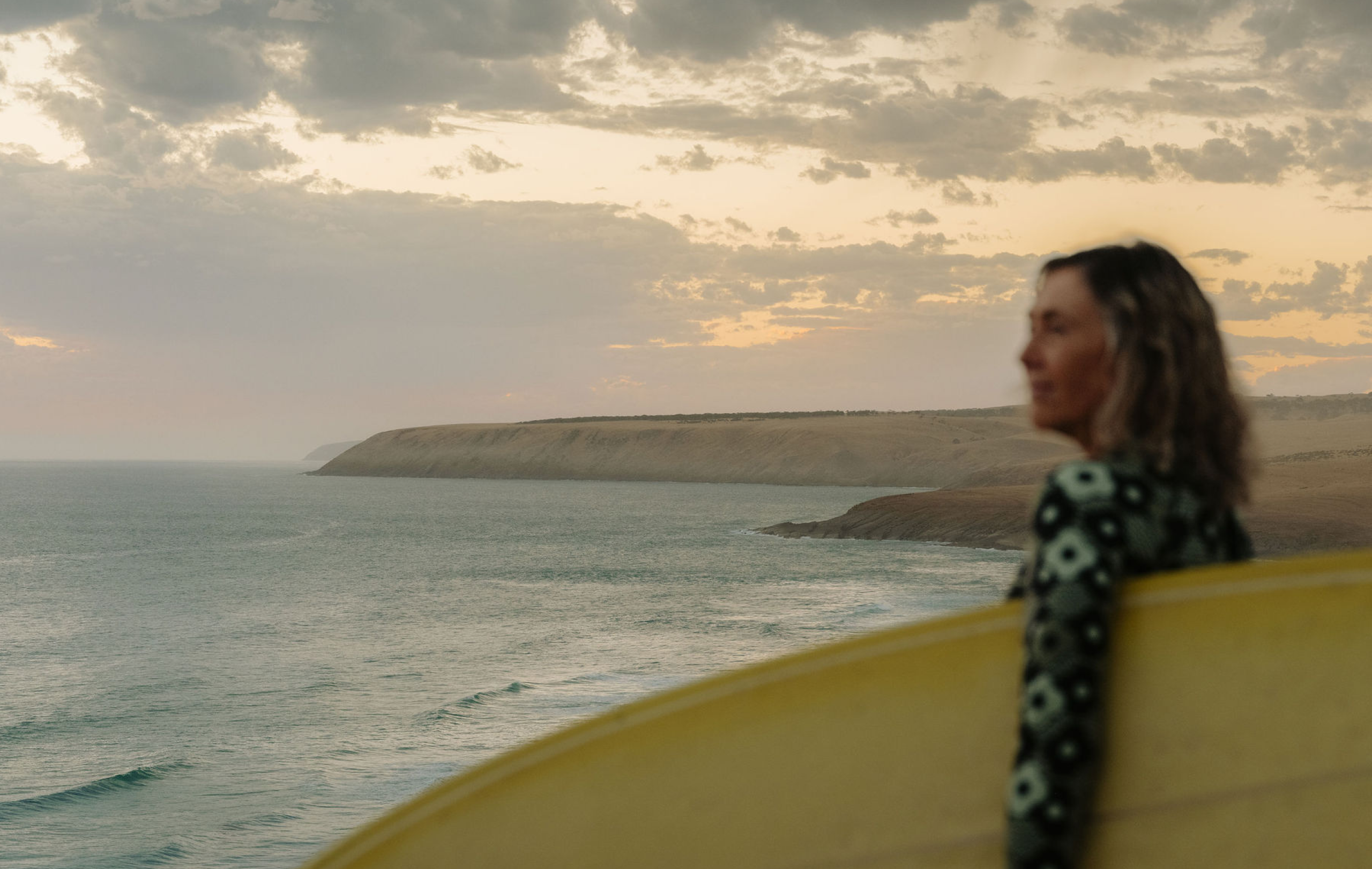 Surfer carrying a yellow surfboard beside the South Australian coast at sunset.