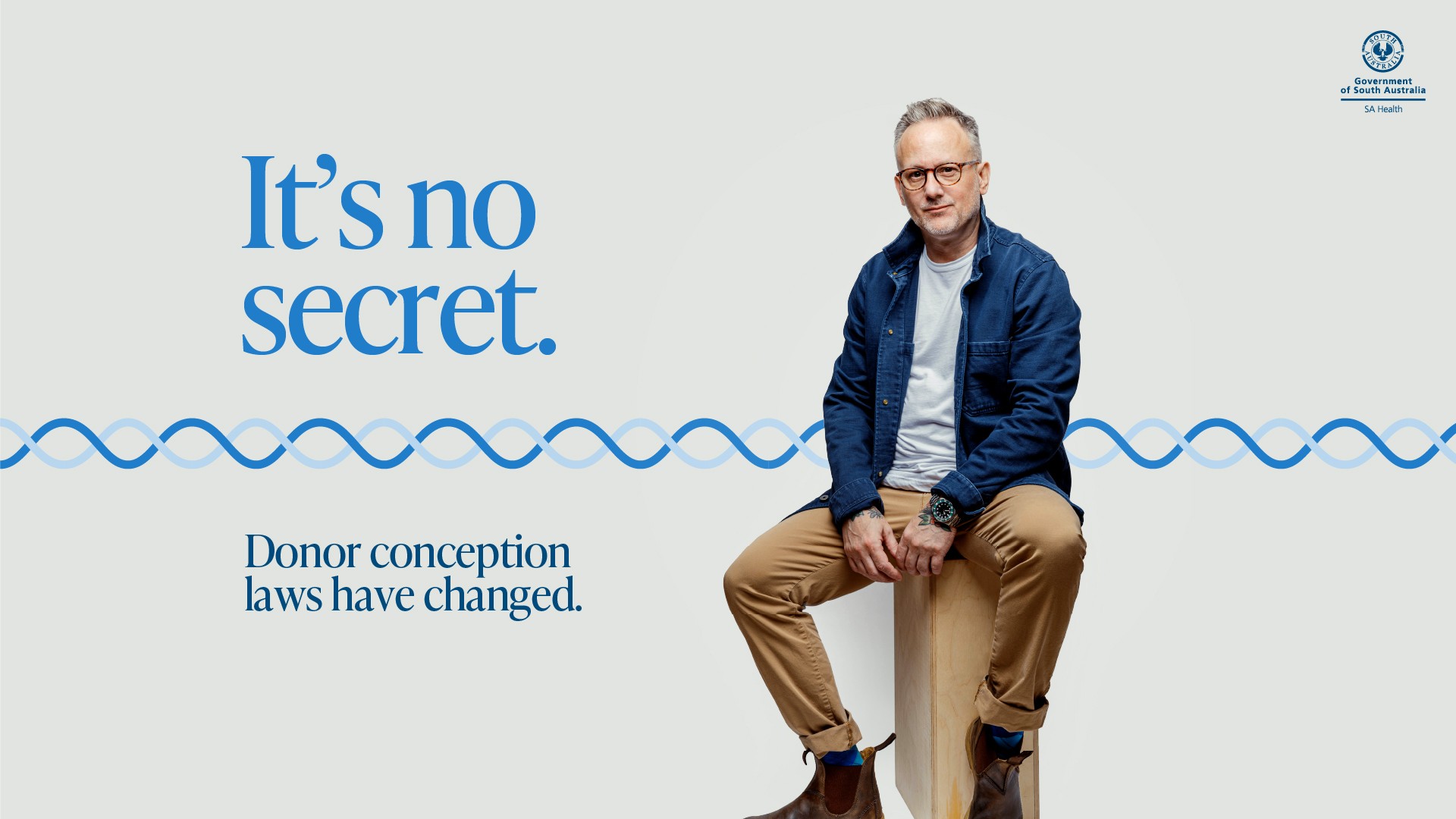 An SA Health public message featuring a person with glasses, a blue overshirt, white t-shirt, beige chinos, and brown leather Blundstone boots sitting on a wooden block against a plain light grey background. Large blue text on the left reads "It's no secret. Donor conception laws have changed.", with a blue wave line design pattern running horizontally across the middle. The Government of South Australia SA Health logo is displayed in the top right corner.