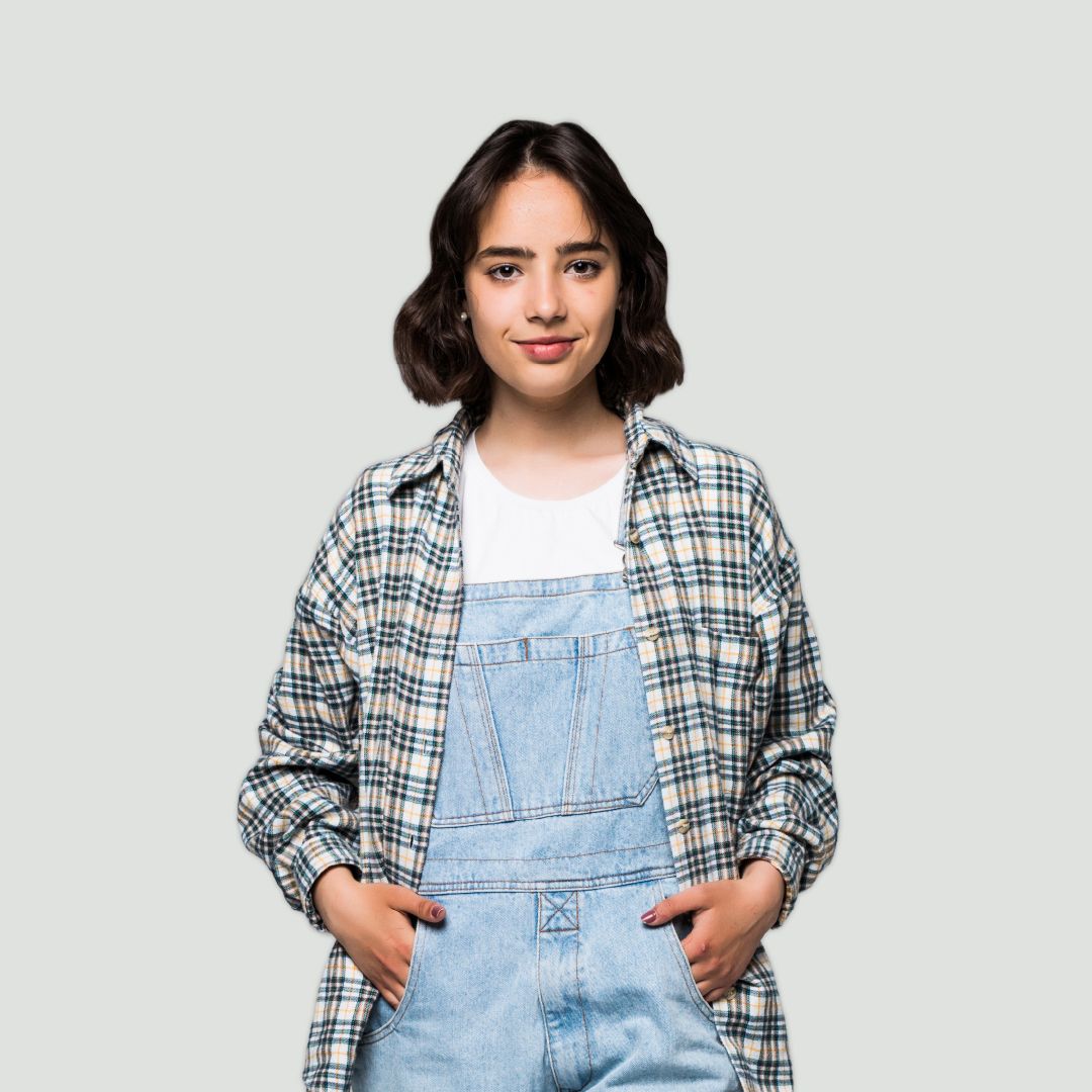 A studio portrait of a person with dark, shoulder-length wavy hair against a plain grey background. The person wears light blue denim dungarees over a white t-shirt, topped with an open plaid flannel shirt, standing with hands in front pockets and looking directly at the camera with a slight smile.