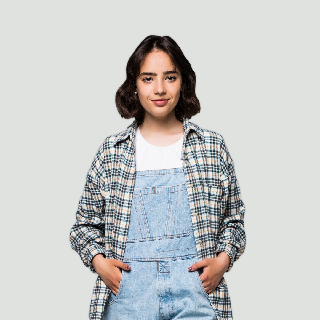 A studio portrait of a person with dark, shoulder-length wavy hair against a plain grey background. The person wears light blue denim dungarees over a white t-shirt, topped with an open plaid flannel shirt, standing with hands in front pockets and looking directly at the camera with a slight smile.