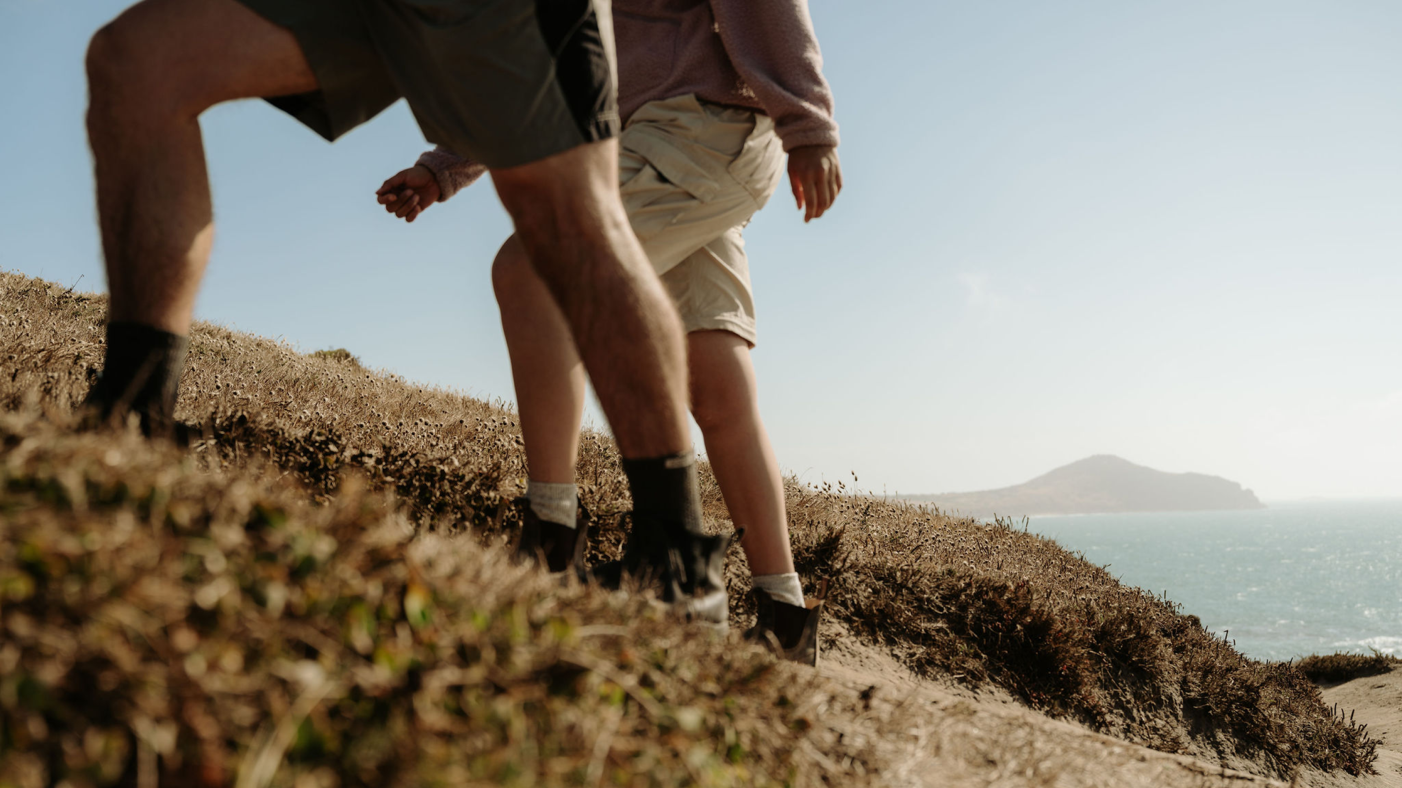 Two people walking through coastal scrub during filming for “The Coast is Calling” campaign.