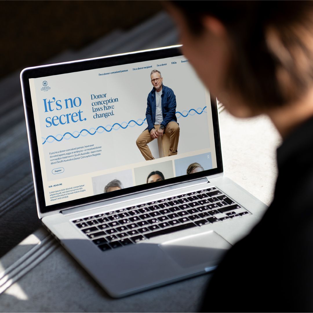 An over-the-shoulder view of a person looking at an open laptop displaying the SA Health "It's no secret. Donor conception laws have changed" website. The screen shows the campaign banner featuring a person with glasses sitting on a wooden block against a blue wave design, alongside navigation options such as "I'm a donor-conceived person", "I'm a donor recipient", and "I'm a donor".