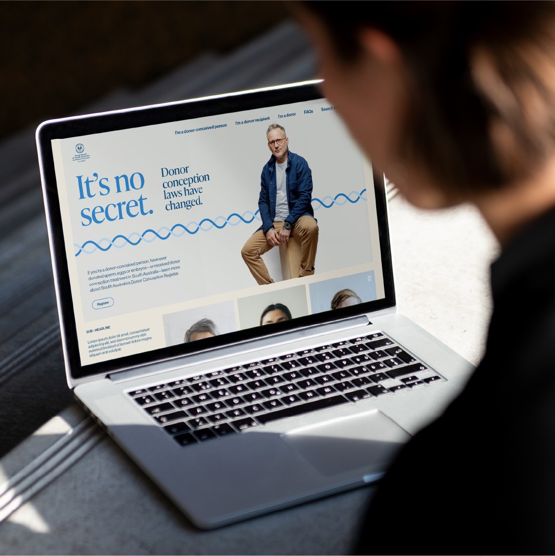 An over-the-shoulder view of a person looking at an open laptop displaying the SA Health "It's no secret. Donor conception laws have changed" website. The screen shows the campaign banner featuring a person with glasses sitting on a wooden block against a blue wave design, alongside navigation options such as "I'm a donor-conceived person", "I'm a donor recipient", and "I'm a donor".