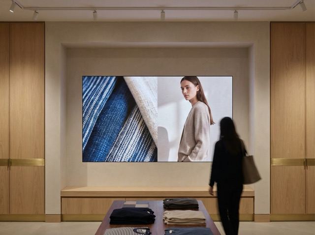 Modern retail store interior with wooden shelves and a large digital screen displaying a close-up of fabric and a model wearing a beige sweater.