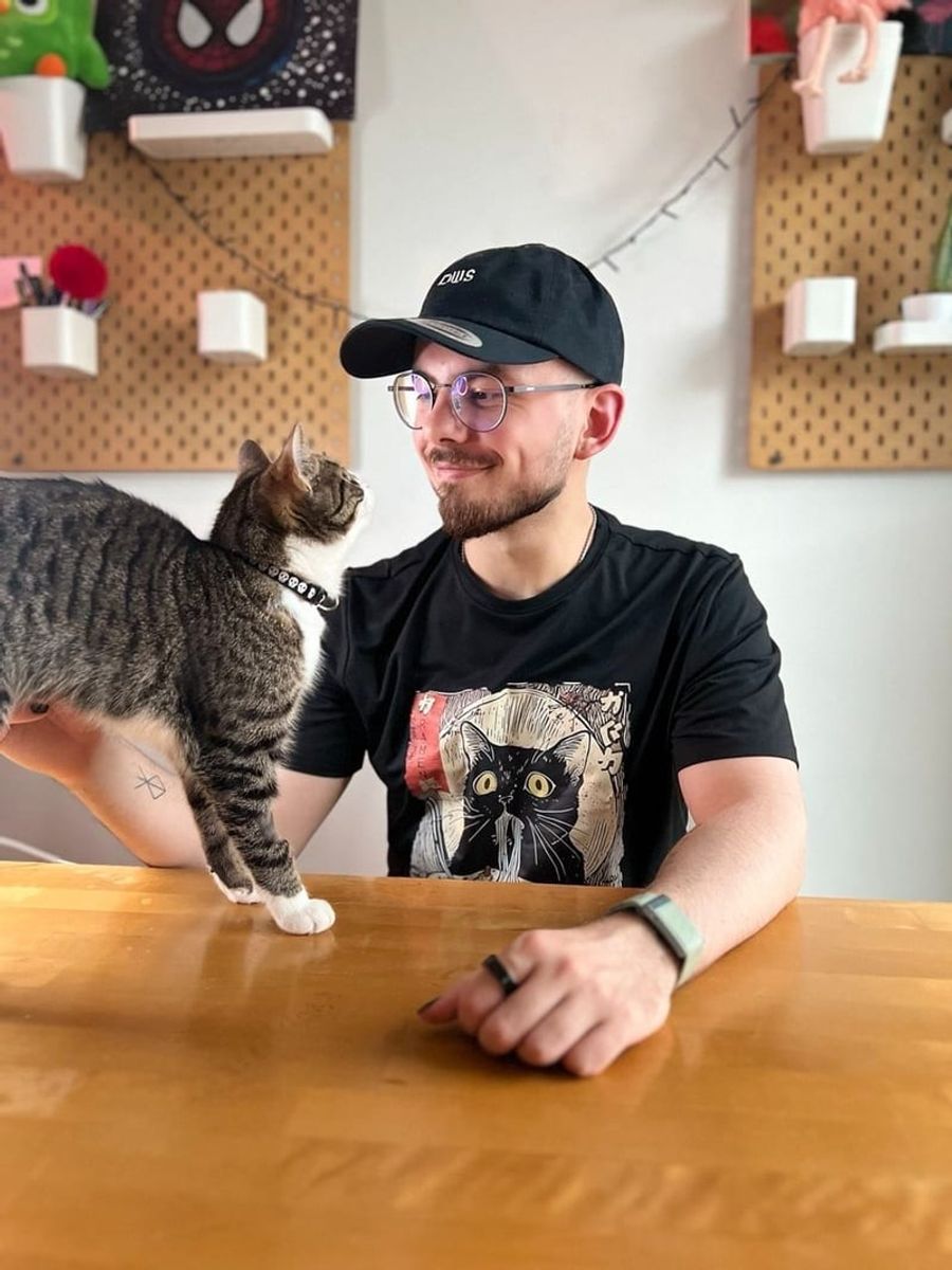 A man in a cap and glasses at a desk, turning to a cat standing on the table