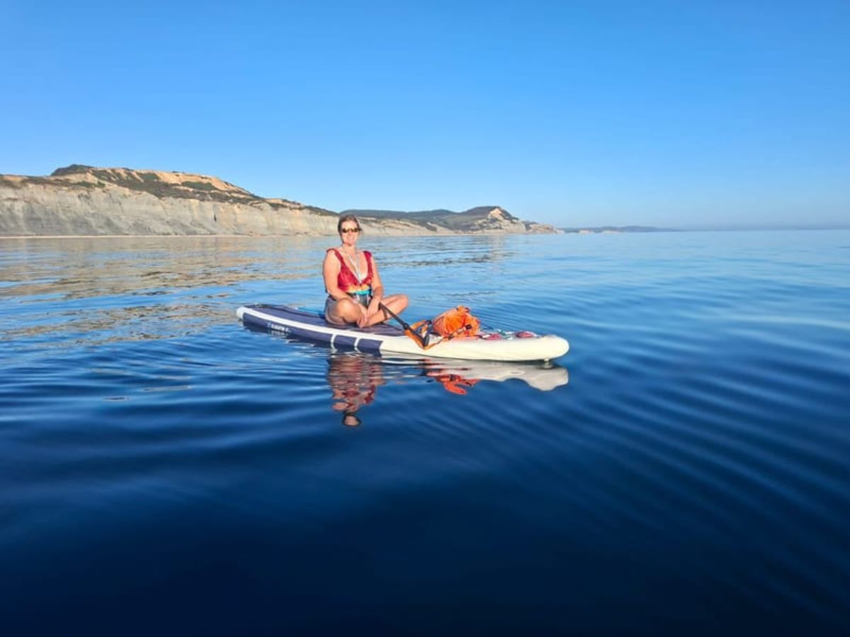 A paddleboarder sitting on flat blue sea beneath a cliff