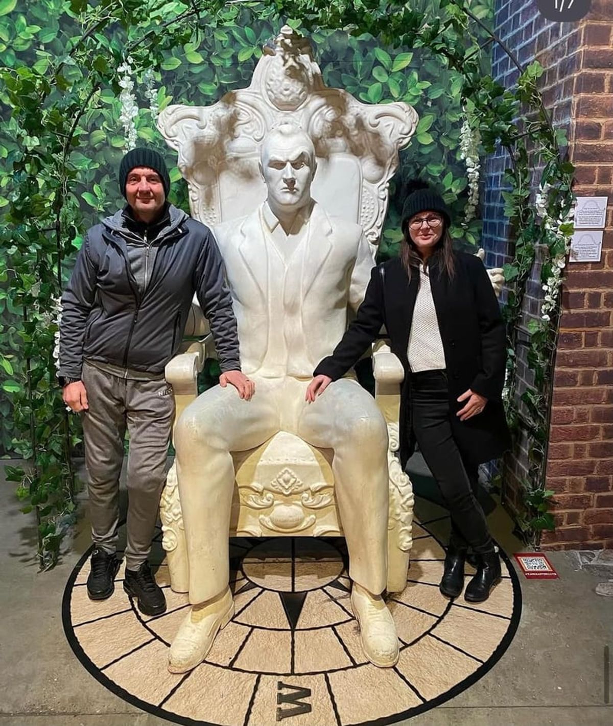 Two people standing either side of a large stone sculpture of a seated figure