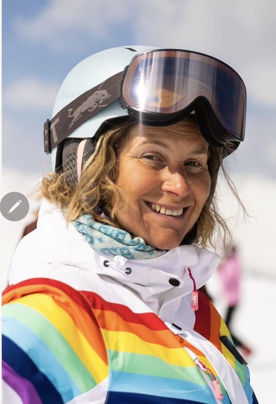 Cath on a ski slope in a rainbow ski jacket, goggles pushed up, smiling
