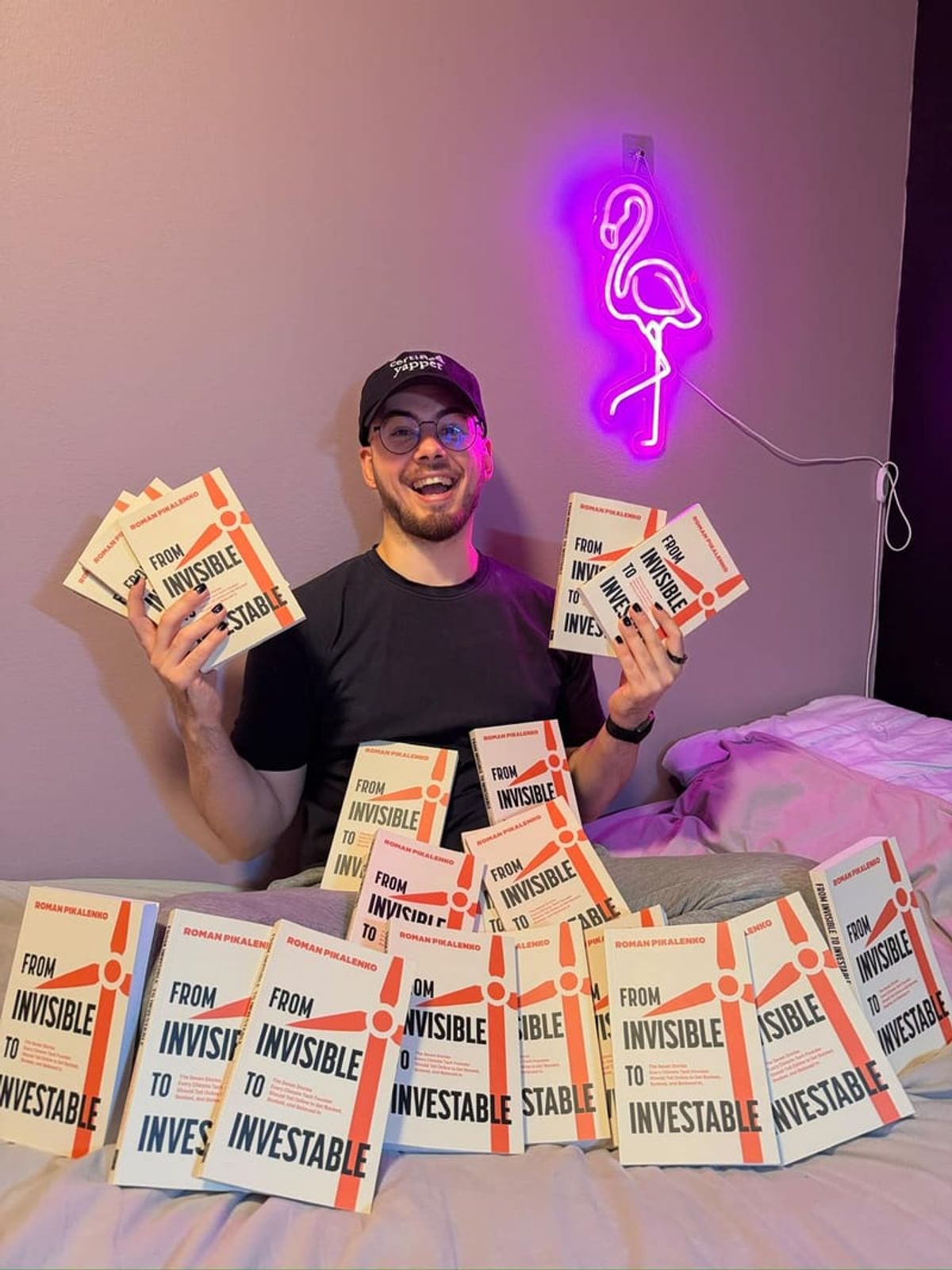 A man surrounded by copies of his own published book, holding up a handful of them