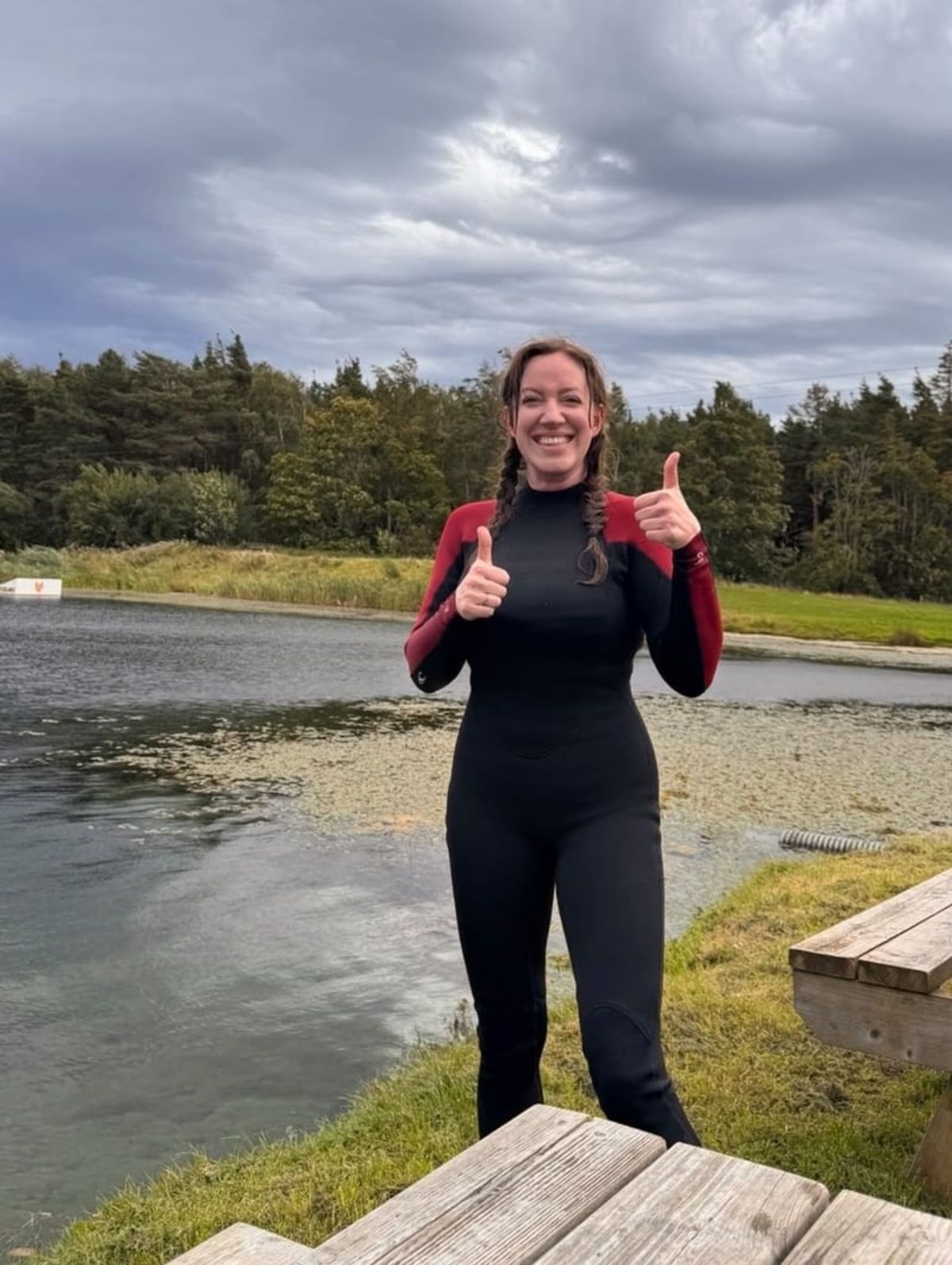 A wetsuited figure giving two thumbs up beside a lake