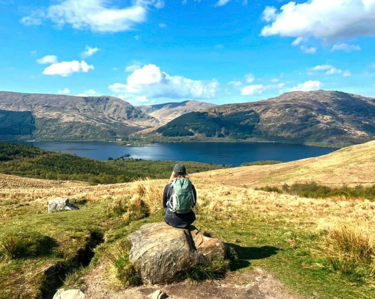 A walker sitting on a rock above a loch with mountains beyond