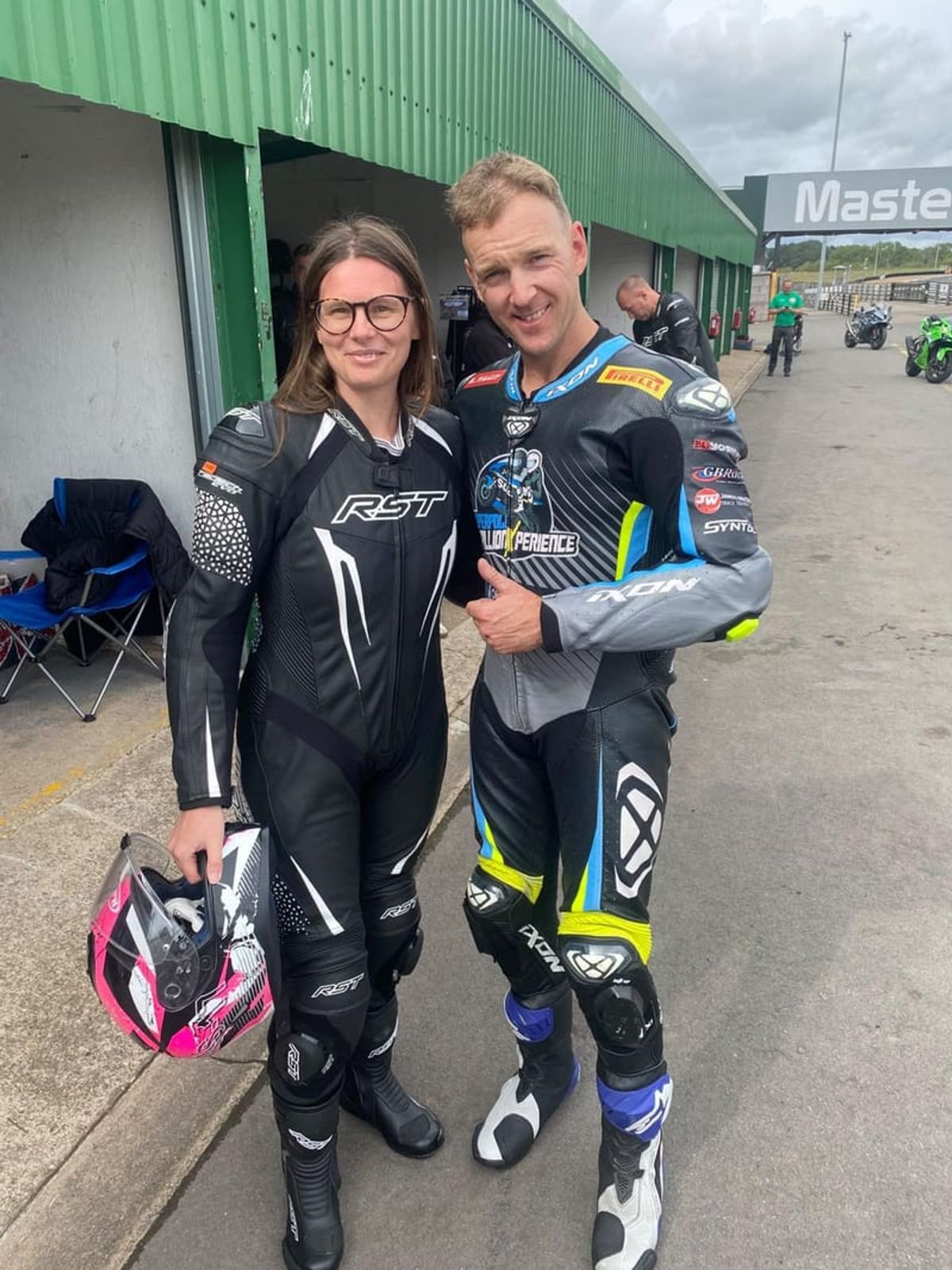 Two people in motorcycle racing suits standing together in a pit lane