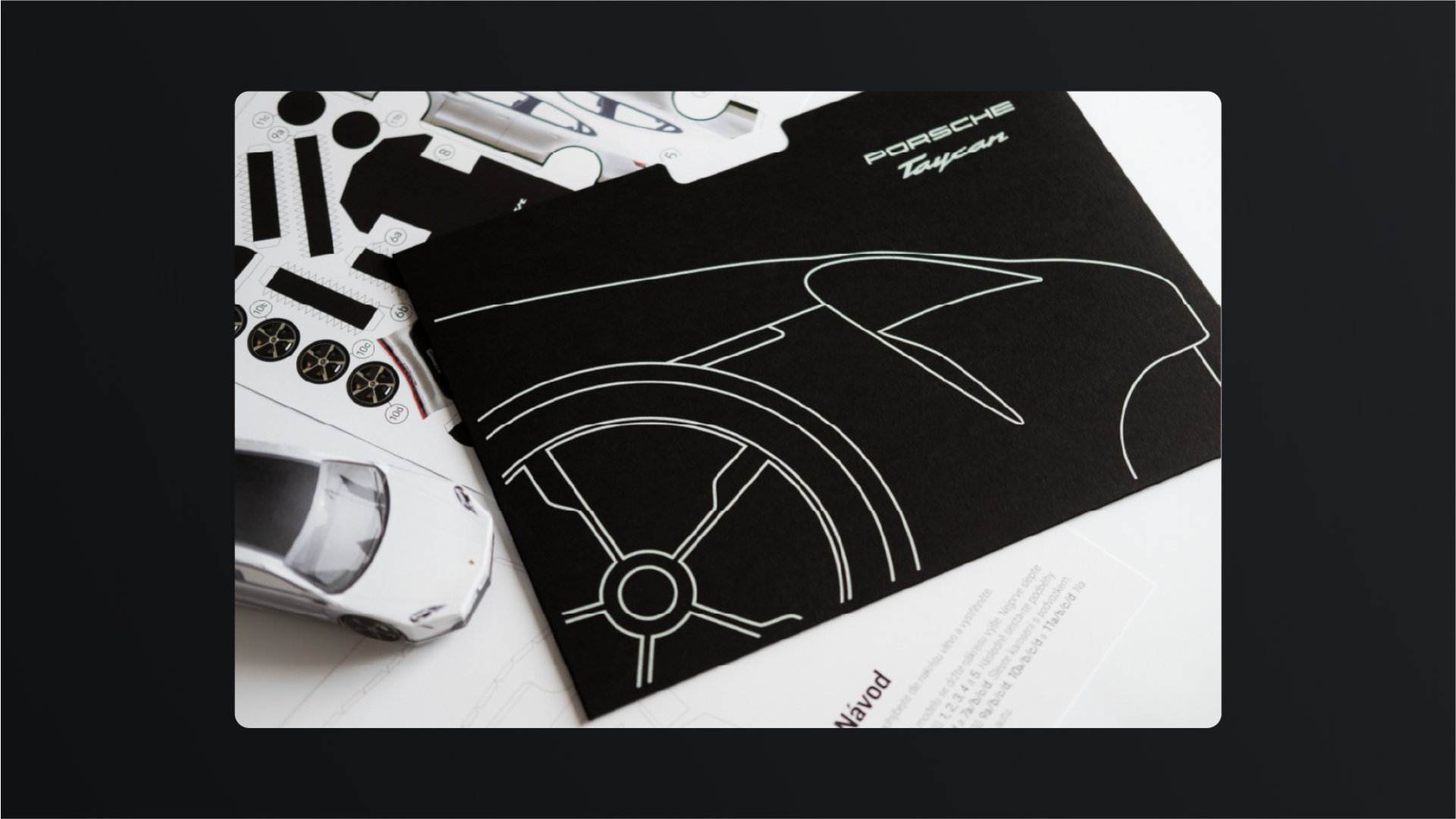 A black Porsche Taycan folder with a line drawing of the car, a paper model and instructions