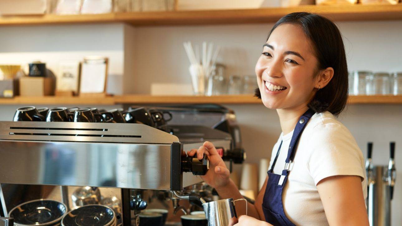Lachende barista in blauw schort bedient de espressomachine achter de bar van een koffiezaak