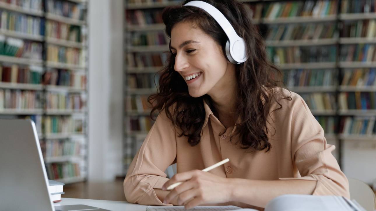 Lachende vrouw met koptelefoon werkt aan een laptop voor een grote boekenkast in een bibliotheek