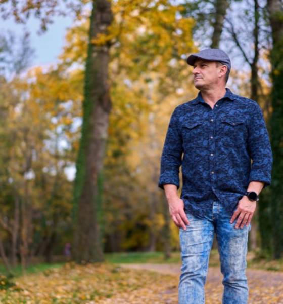 Man met pet loopt over een bospad vol herfstbladeren en kijkt opzij