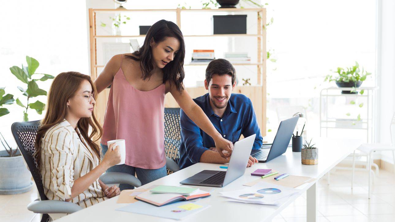 Drie collega's overleggen aan een tafel met laptops, een van hen wijst iets aan op een document