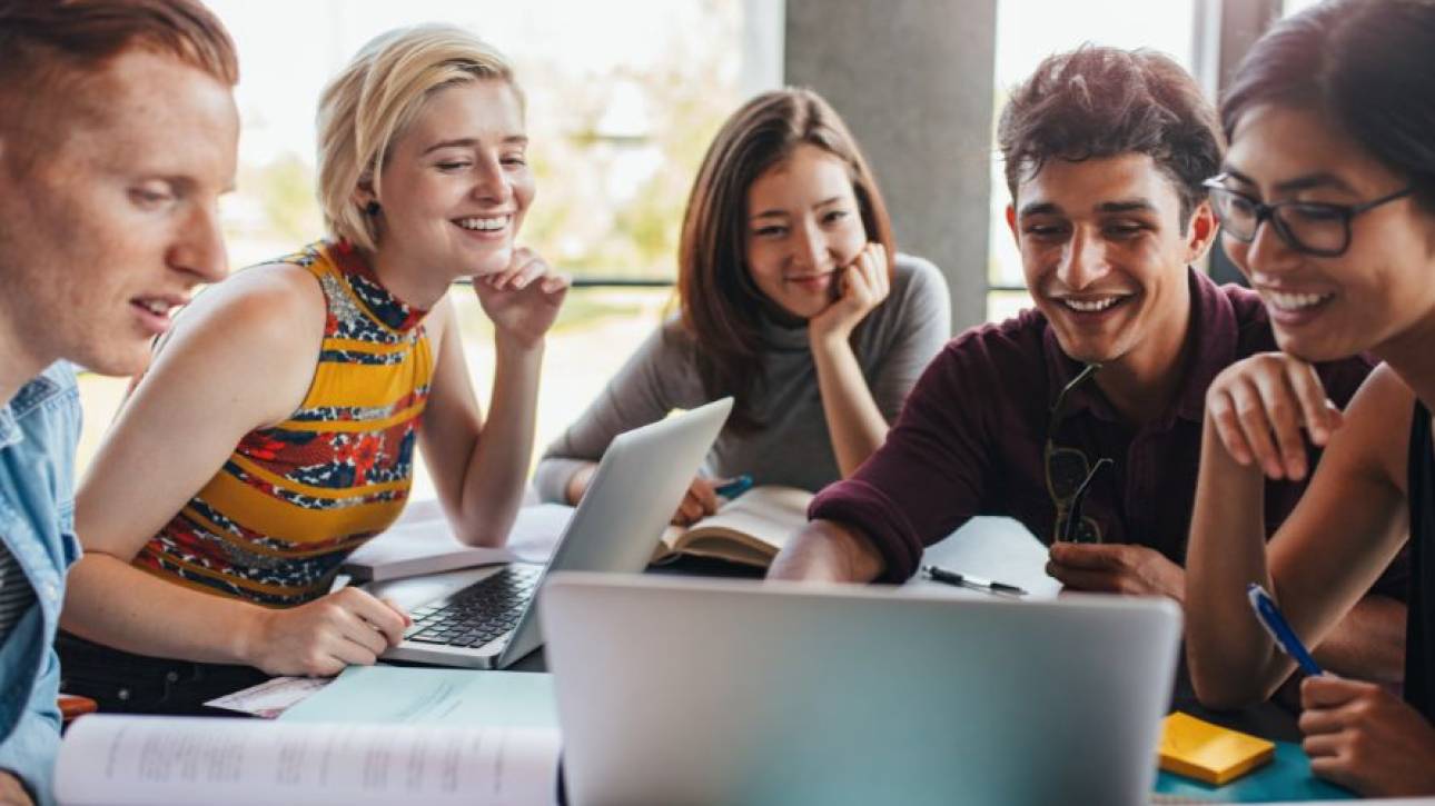 Vijf jonge mensen lachen samen achter laptops, boeken en aantekeningen aan een tafel