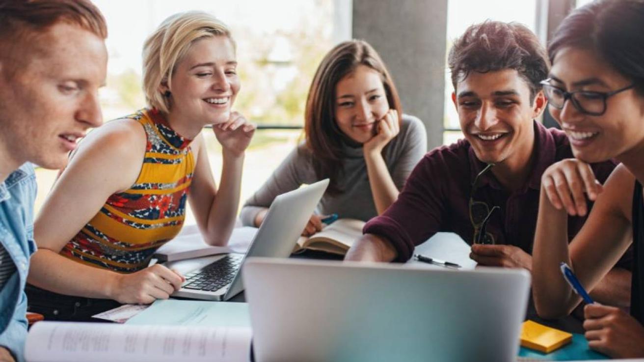Vijf jonge mensen lachen samen achter laptops, boeken en aantekeningen aan een tafel