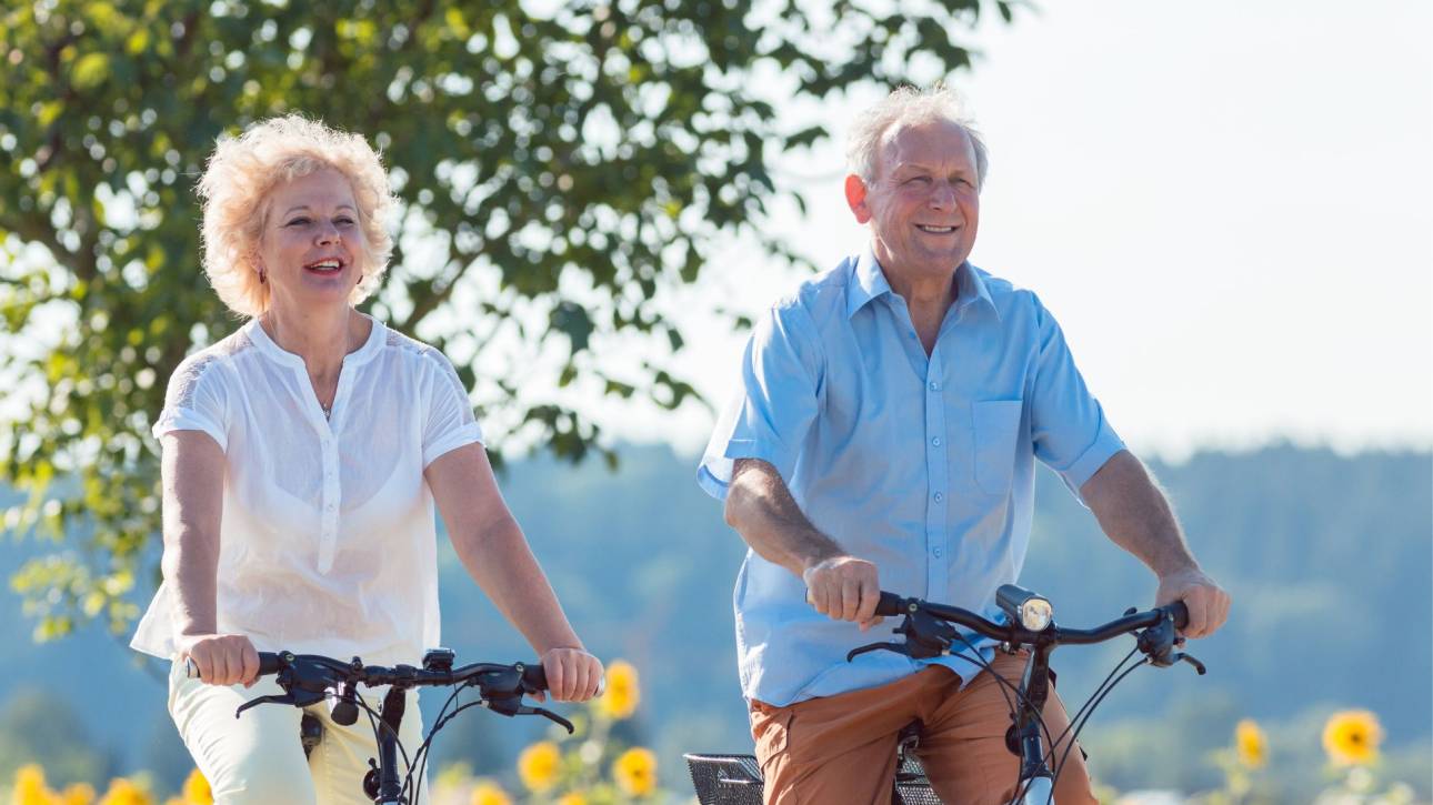 Twee oudere personen fietsen lachend langs een veld met zonnebloemen in de zon