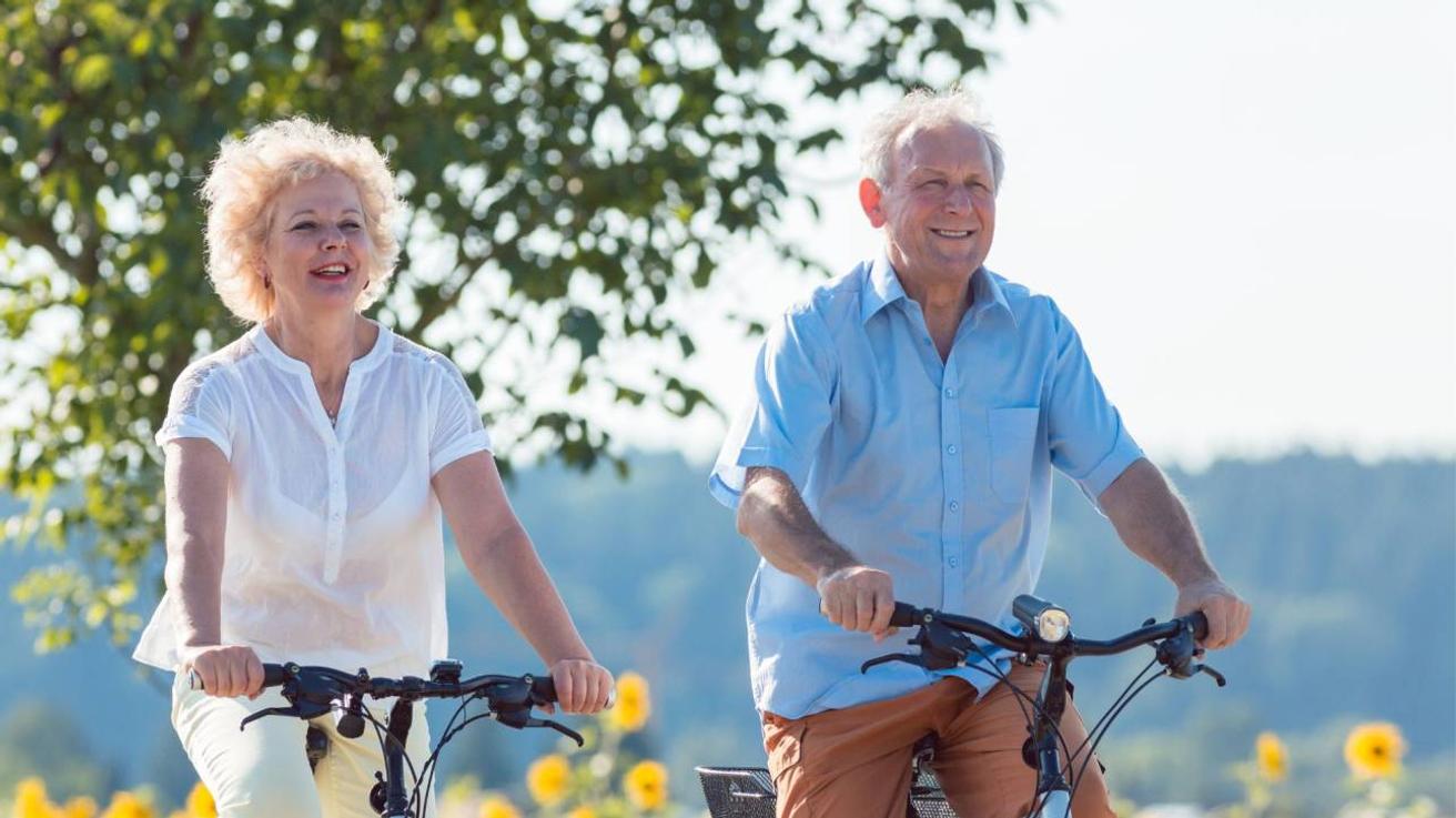 Twee oudere personen fietsen lachend langs een veld met zonnebloemen in de zon