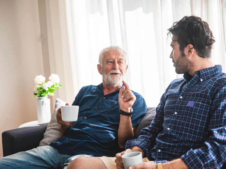 Oudere man met grijze baard en jongere man drinken koffie en praten met elkaar op de bank