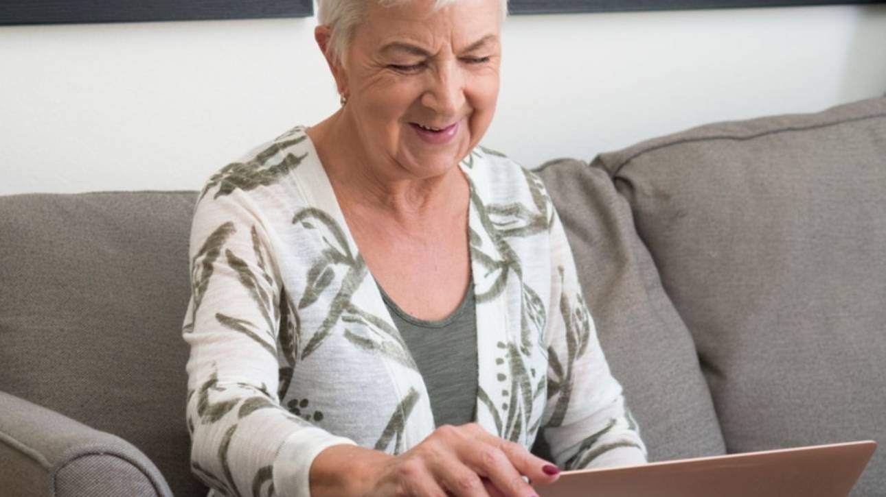 Vrouw met wit haar zit lachend op de bank en typt op een laptop