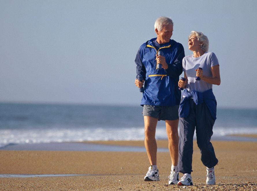 Twee oudere mensen joggen lachend naast elkaar over het strand langs de zee