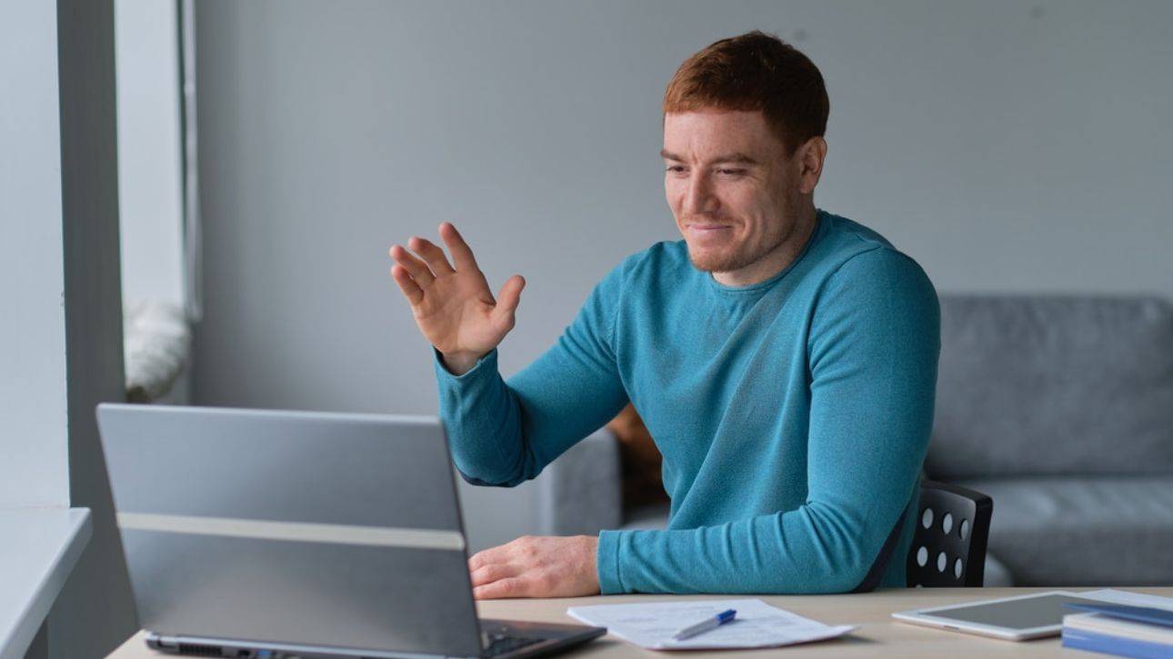 Man in blauwe trui zwaait naar het scherm van zijn laptop tijdens een videogesprek aan een bureau