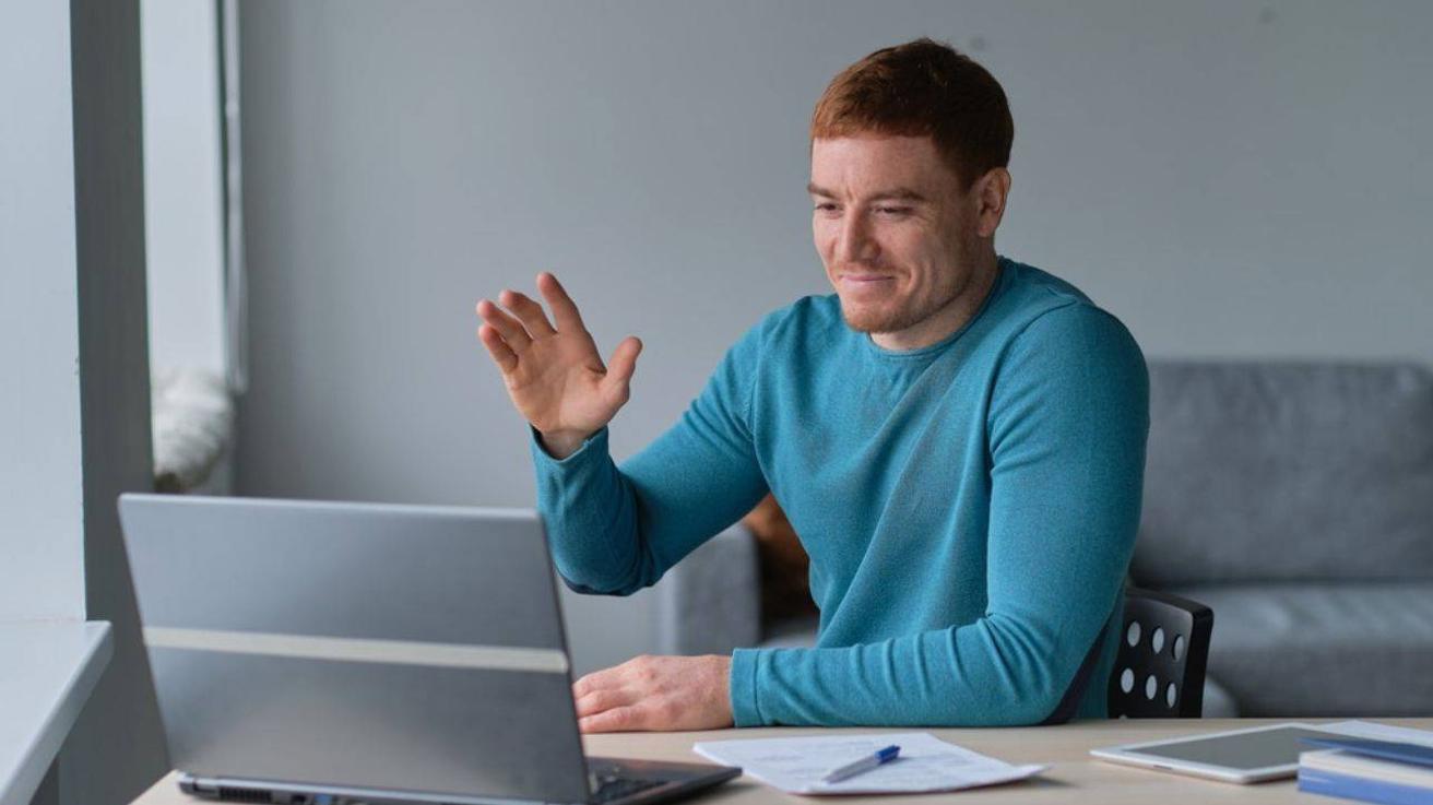 Man in blauwe trui zwaait naar het scherm van zijn laptop tijdens een videogesprek aan een bureau
