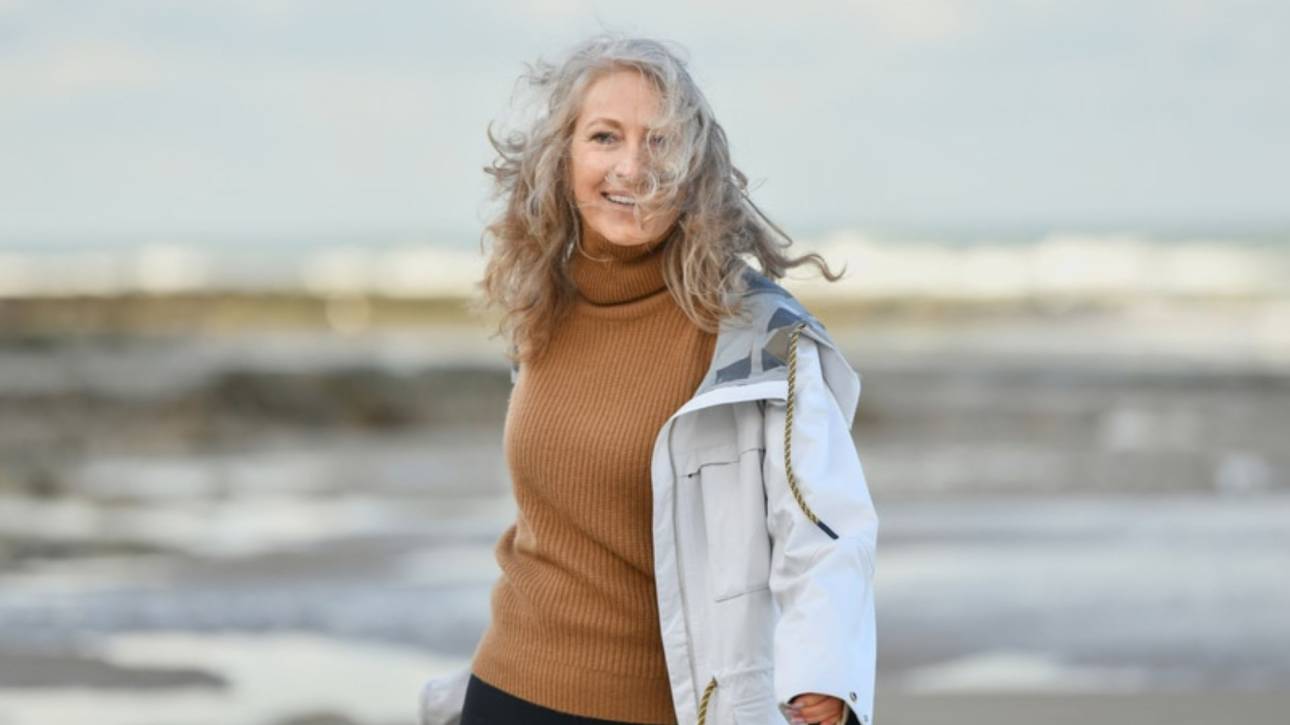 Vrouw met grijze krullen in coltrui en jas loopt glimlachend over een winderig strand