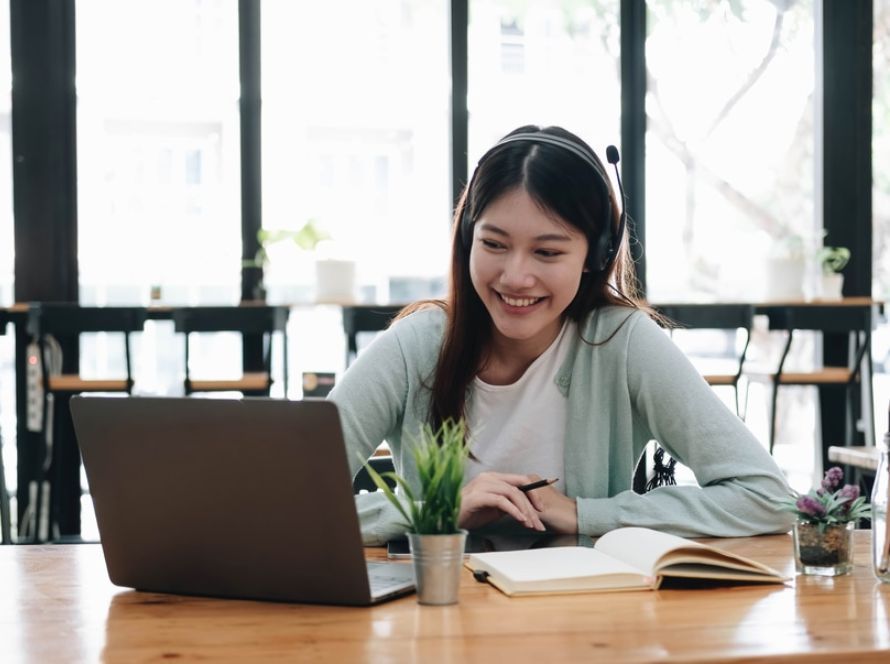 Lachende vrouw met headset werkt aan een laptop met een opengeslagen notitieboek ernaast