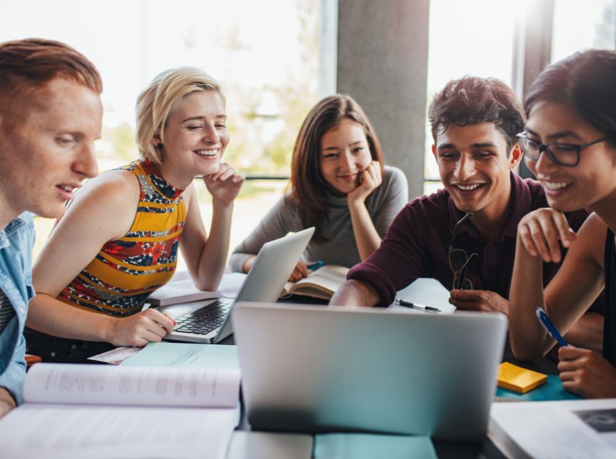 Vijf jonge mensen werken lachend samen achter een laptop, met studieboeken en aantekeningen op tafel