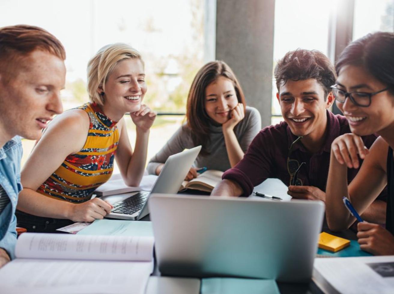 Vijf jonge mensen werken lachend samen achter een laptop, met studieboeken en aantekeningen op tafel