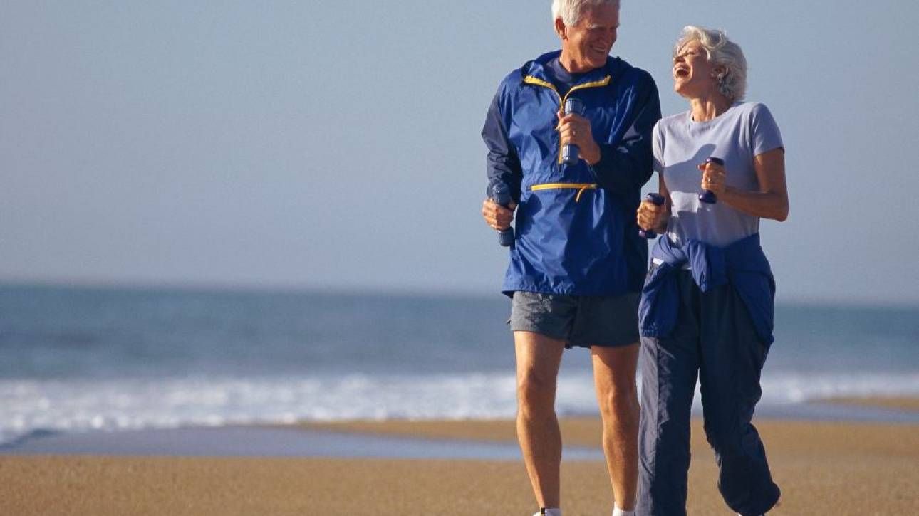 Twee oudere personen joggen lachend naast elkaar over het strand langs de zee