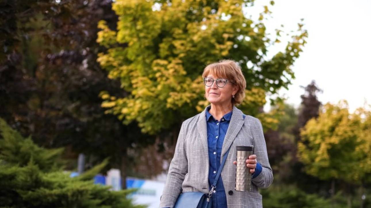 Vrouw met bril en geruit colbert loopt buiten met een thermosbeker en schoudertas langs herfstige bomen