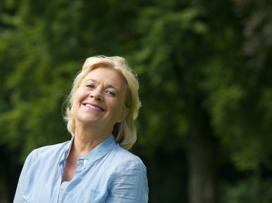 Lachende vrouw met blond haar in een lichtblauwe blouse buiten voor groene bomen