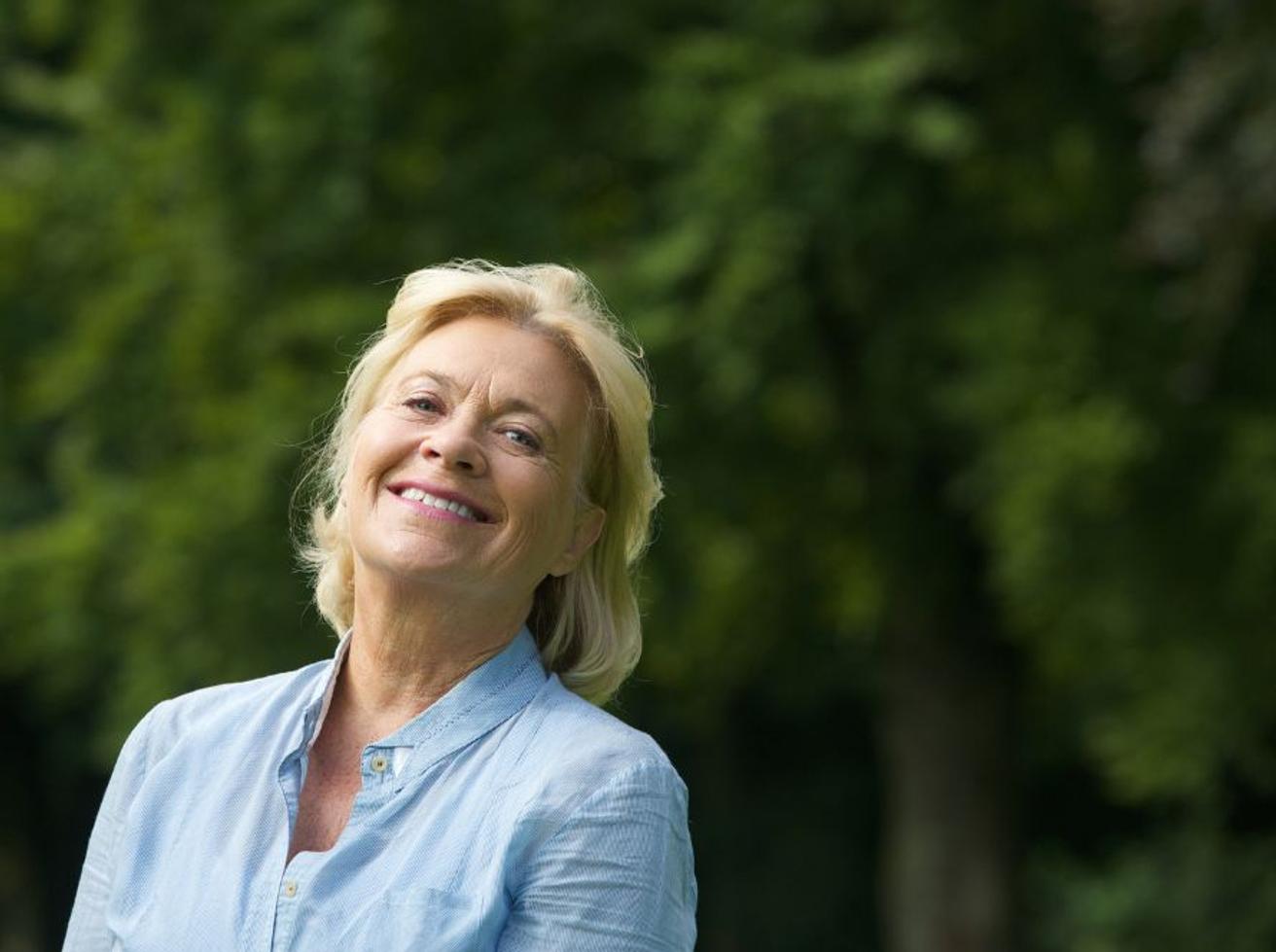 Lachende vrouw met blond haar in een lichtblauwe blouse buiten voor groene bomen