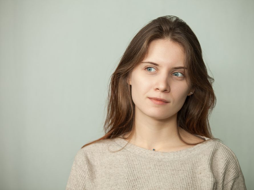 Vrouw met lang bruin haar kijkt peinzend opzij tegen een lichtgrijze achtergrond