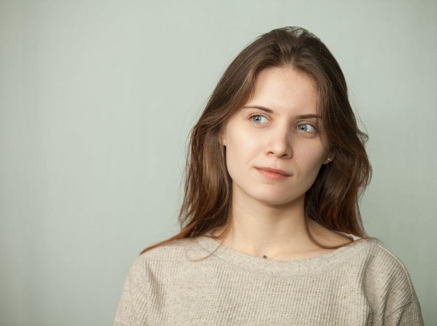 Vrouw met lang bruin haar kijkt peinzend opzij tegen een lichtgrijze achtergrond