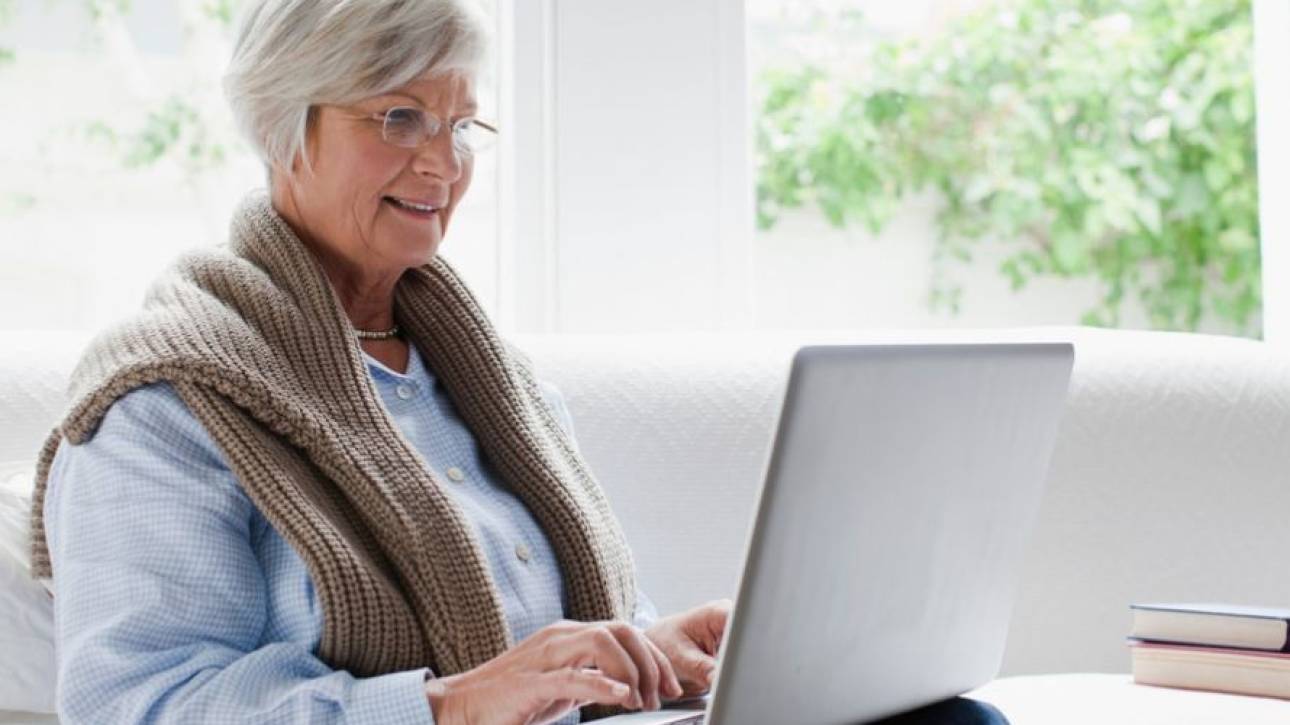 Vrouw met kort grijs haar en bril typt glimlachend op een laptop op de bank, met een stapel boeken ernaast