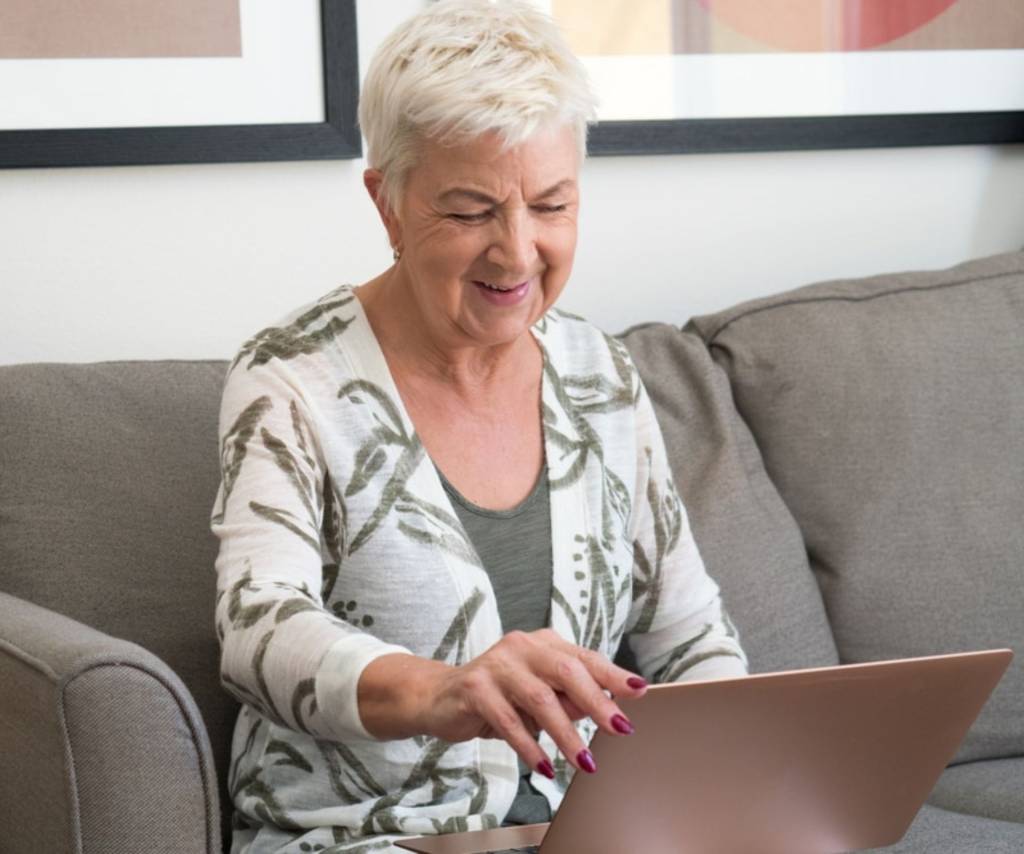 Oudere persoon typt lachend op een laptop op de bank in de woonkamer