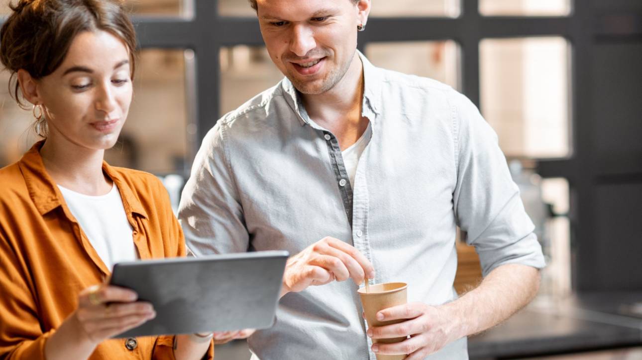 Twee collega's kijken samen naar een tablet, een van hen houdt een koffiebeker vast