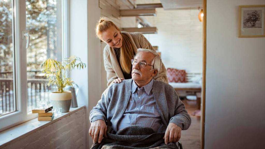 Jonge vrouw staat glimlachend achter een oudere man in een rolstoel bij een raam in huis